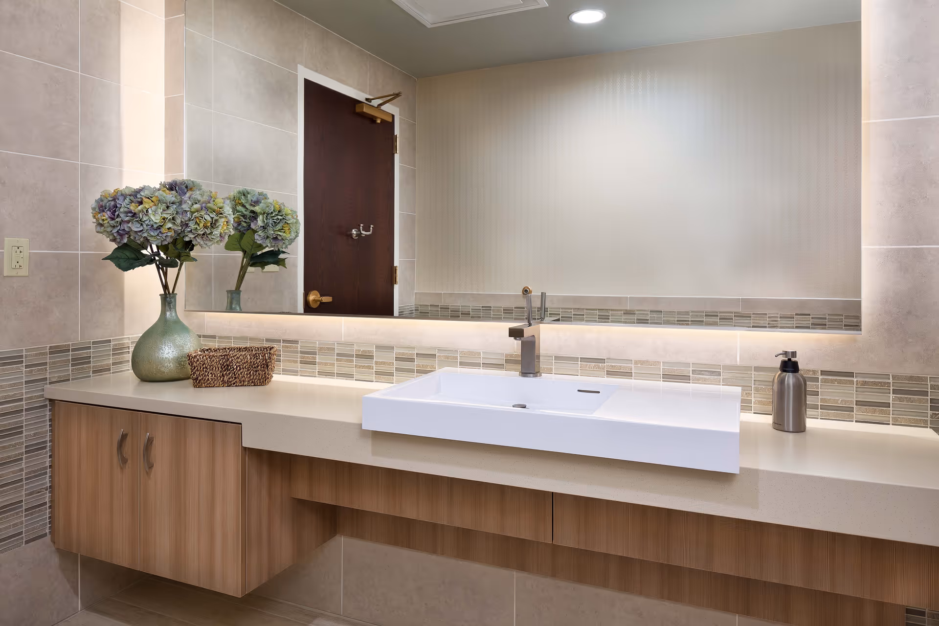 A modern bathroom sink area with a rectangular white basin, a sleek faucet, a large mirror above the sink, a vase with purple and green flowers, a small woven basket, and a soap dispenser on a beige countertop with wooden cabinets below.