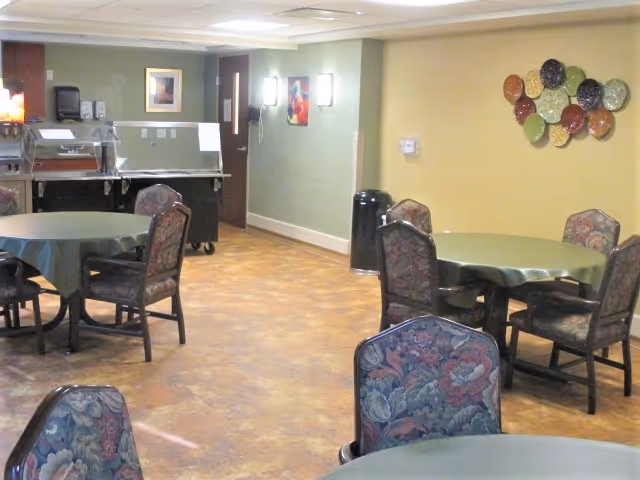 Dining area with round tables covered in green tablecloths, floral-upholstered chairs, a serving cart, and decorative plates on the wall.