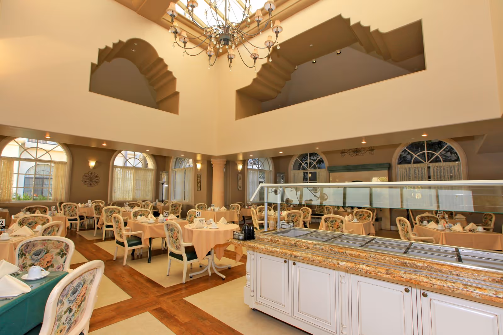 A spacious dining room with multiple tables covered in peach tablecloths and set with white napkins and cups. The chairs have floral upholstery. The room features large arched windows with sheer curtains, a high ceiling with decorative cutouts, and a chandelier hanging from the center. In the foreground, there is a buffet counter with a glass sneeze guard.