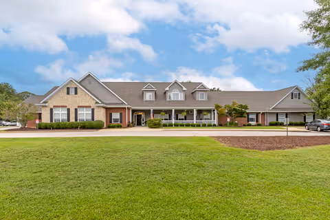 Front exterior view of Sumter Terrace Assisted Living facility showing a single-story building with a combination of brick and stone facade, multiple windows, a covered porch with white railings, and a well-maintained lawn under a partly cloudy sky.