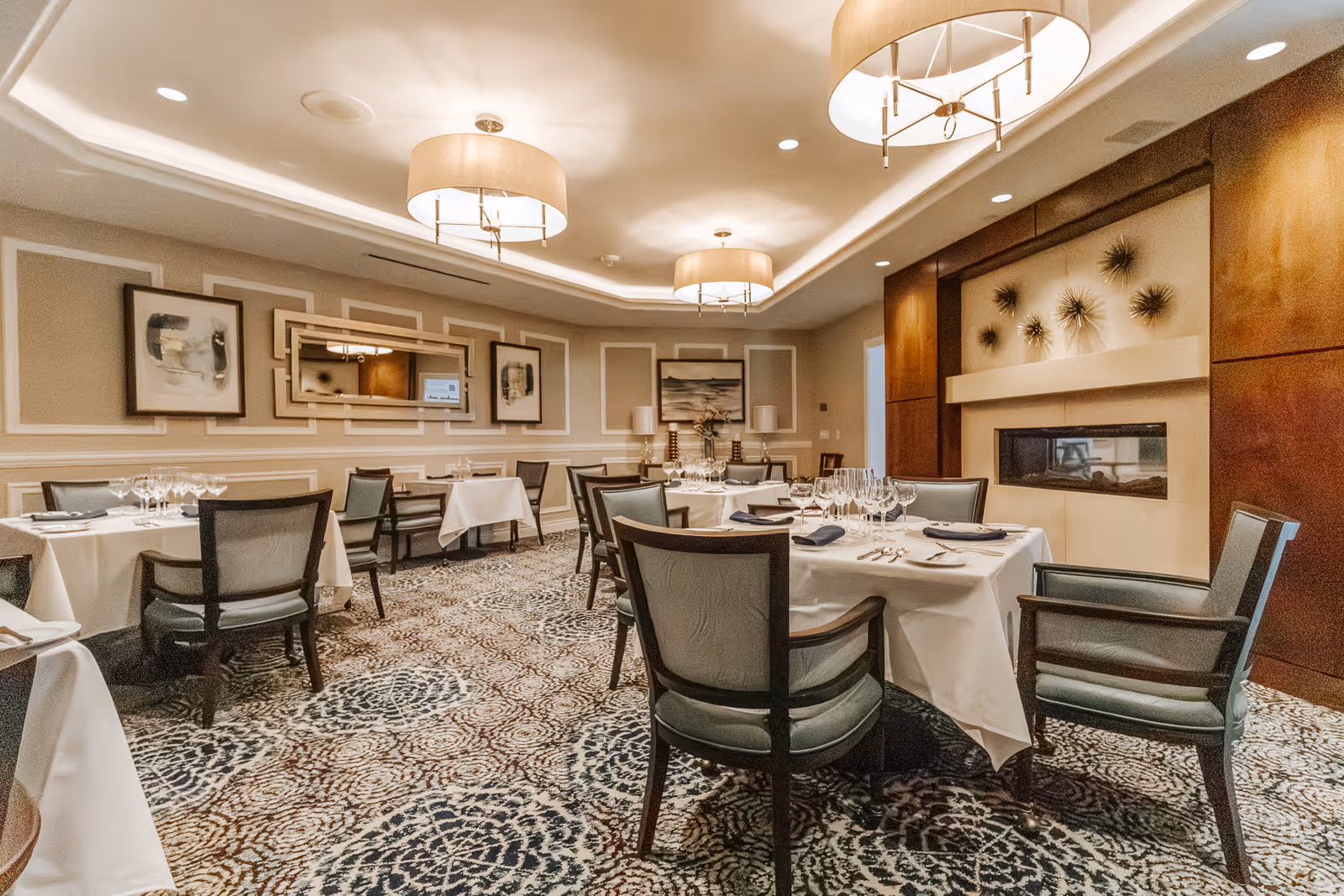 Elegant dining room with multiple set tables, upholstered chairs, chandeliers, decorative wall art, and a fireplace.