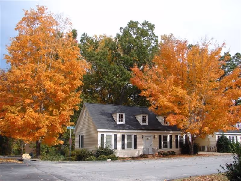 A small beige building with black shutters and a dark roof, surrounded by trees with vibrant orange autumn foliage, situated next to a paved area.