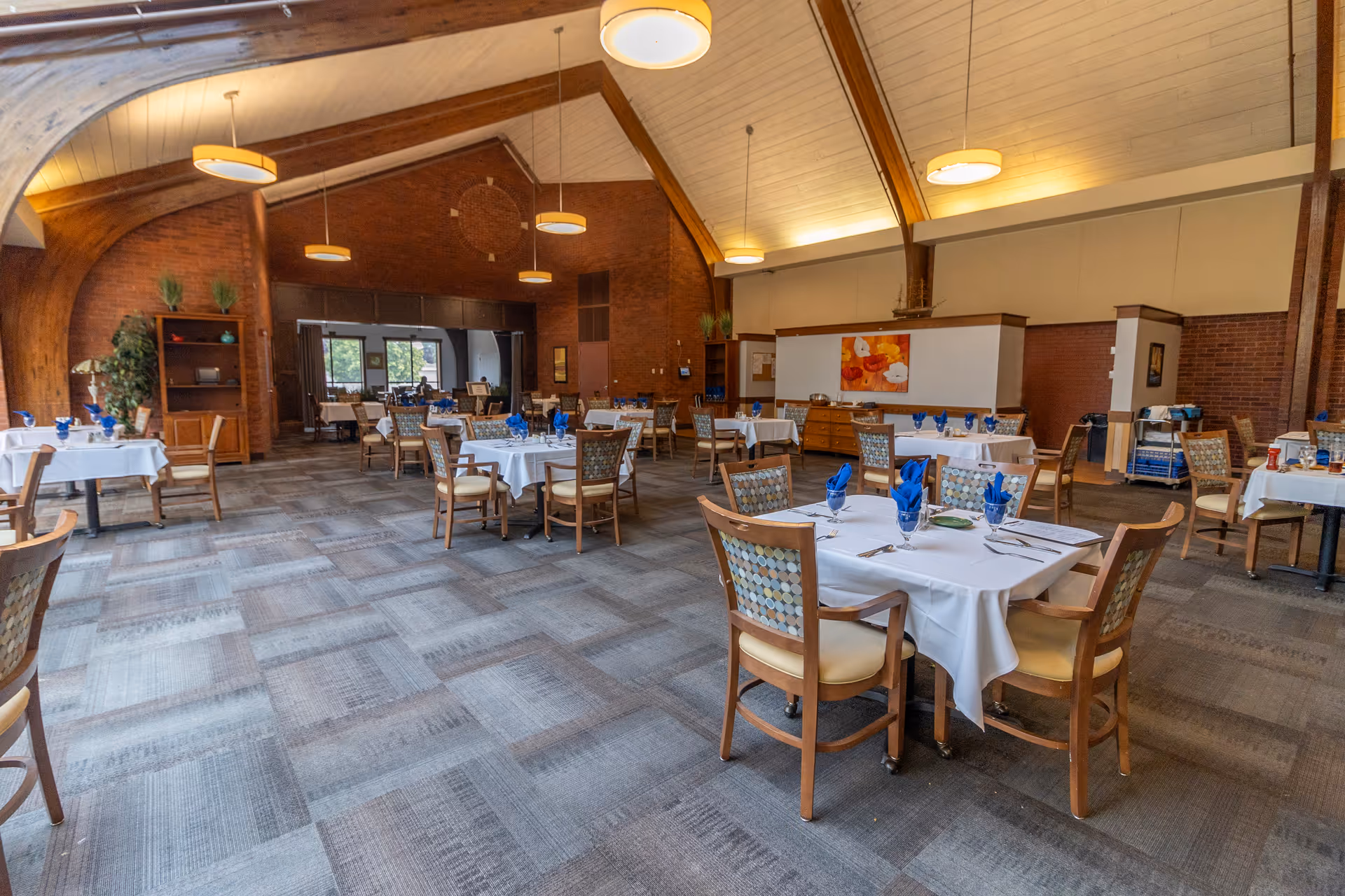 A spacious dining room with multiple tables covered in white tablecloths, each set with blue napkins and glassware. The room features a high vaulted ceiling with wooden beams and round hanging lights. There are patterned chairs around the tables, a carpeted floor, and a brick wall with a colorful painting. Large windows and an open doorway are visible in the background.