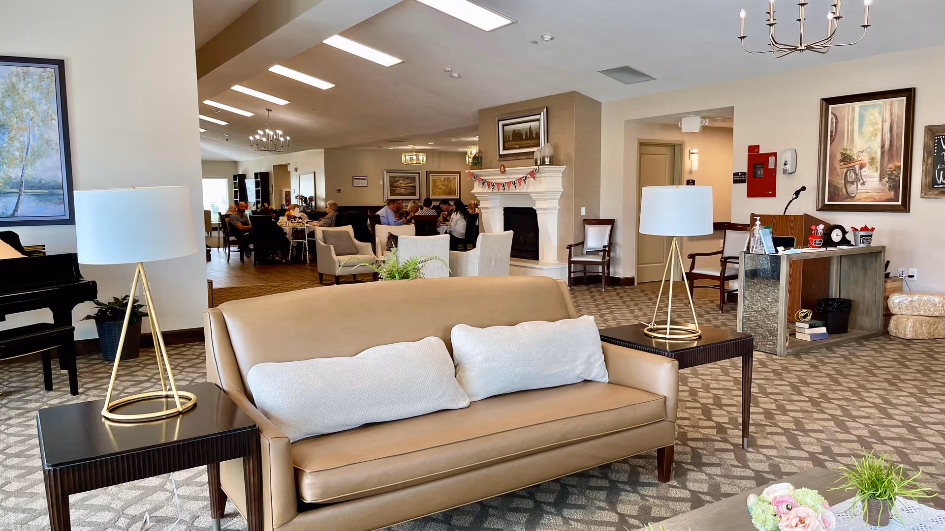 A spacious and well-lit common area in an assisted living facility featuring a tan leather sofa with two white pillows, two side tables each with a modern lamp, a black grand piano, and a fireplace decorated with a garland. In the background, several people are seated at tables, engaged in conversation. The room is decorated with framed artwork and has a patterned carpet.