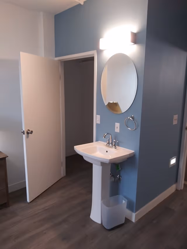 A small bathroom area with a white pedestal sink, a round mirror above it, a wall-mounted light fixture, and a towel ring on a blue wall. The floor is wood-style laminate, and there is an open white door leading to another room. A small trash bin is placed under the sink.