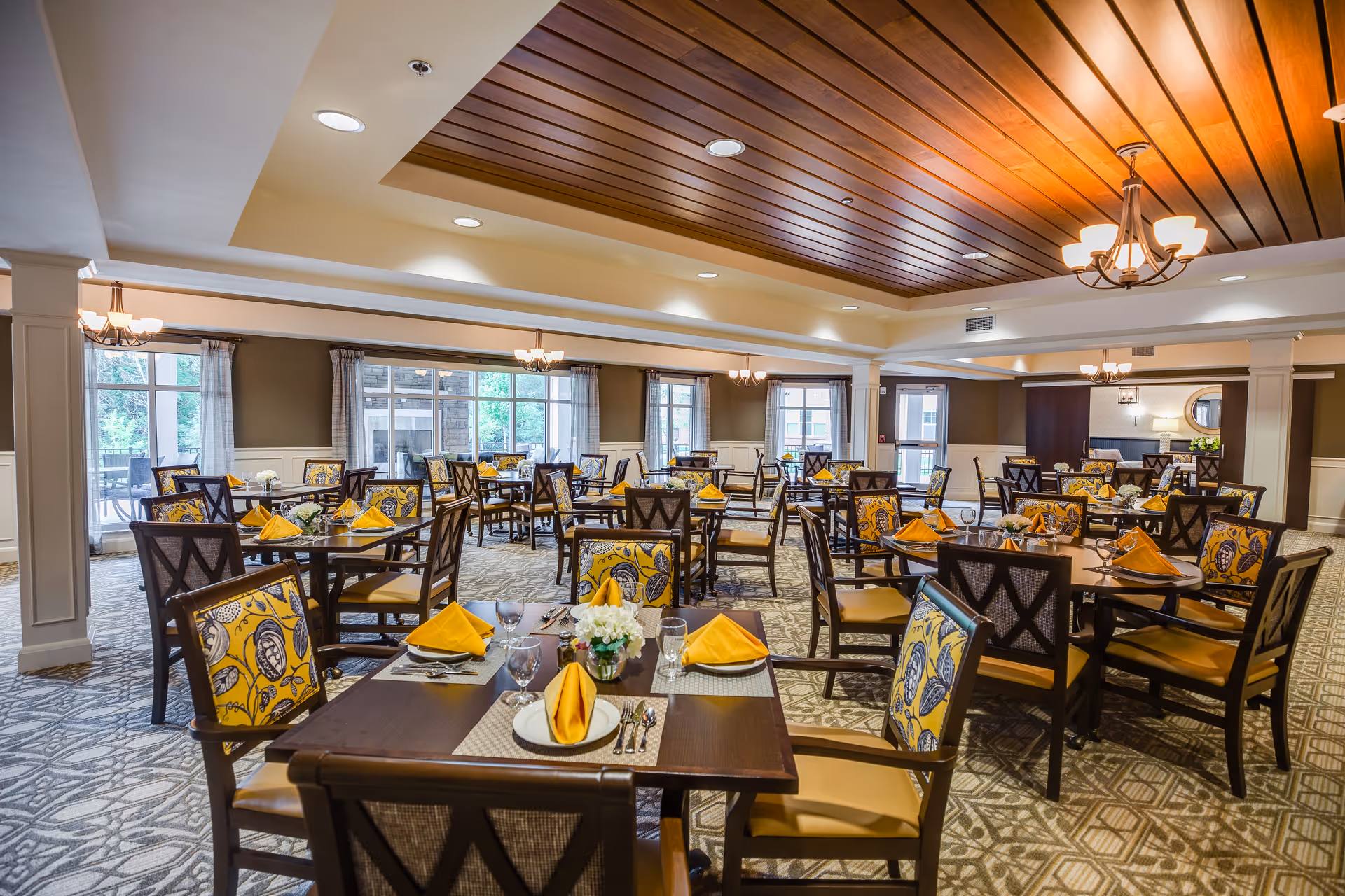 A spacious dining room with multiple dark wood tables and chairs featuring yellow and blue patterned cushions. Each table is set with yellow folded napkins, plates, glasses, and silverware. The room has large windows with sheer curtains allowing natural light to enter, a patterned carpet, and a wooden ceiling with recessed lighting and chandeliers.
