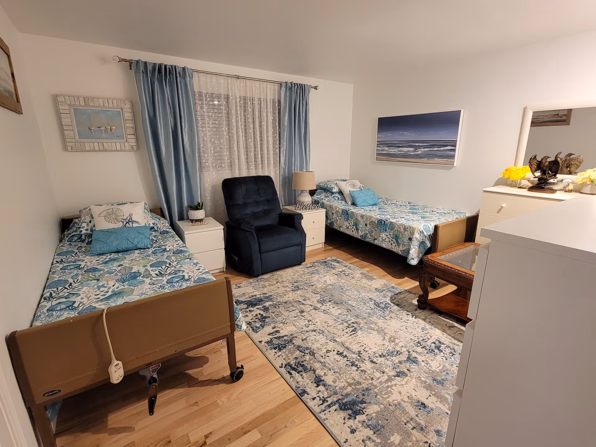 Well-lit bedroom with two twin beds, a navy recliner, nightstands, dresser and coastal-themed decor.