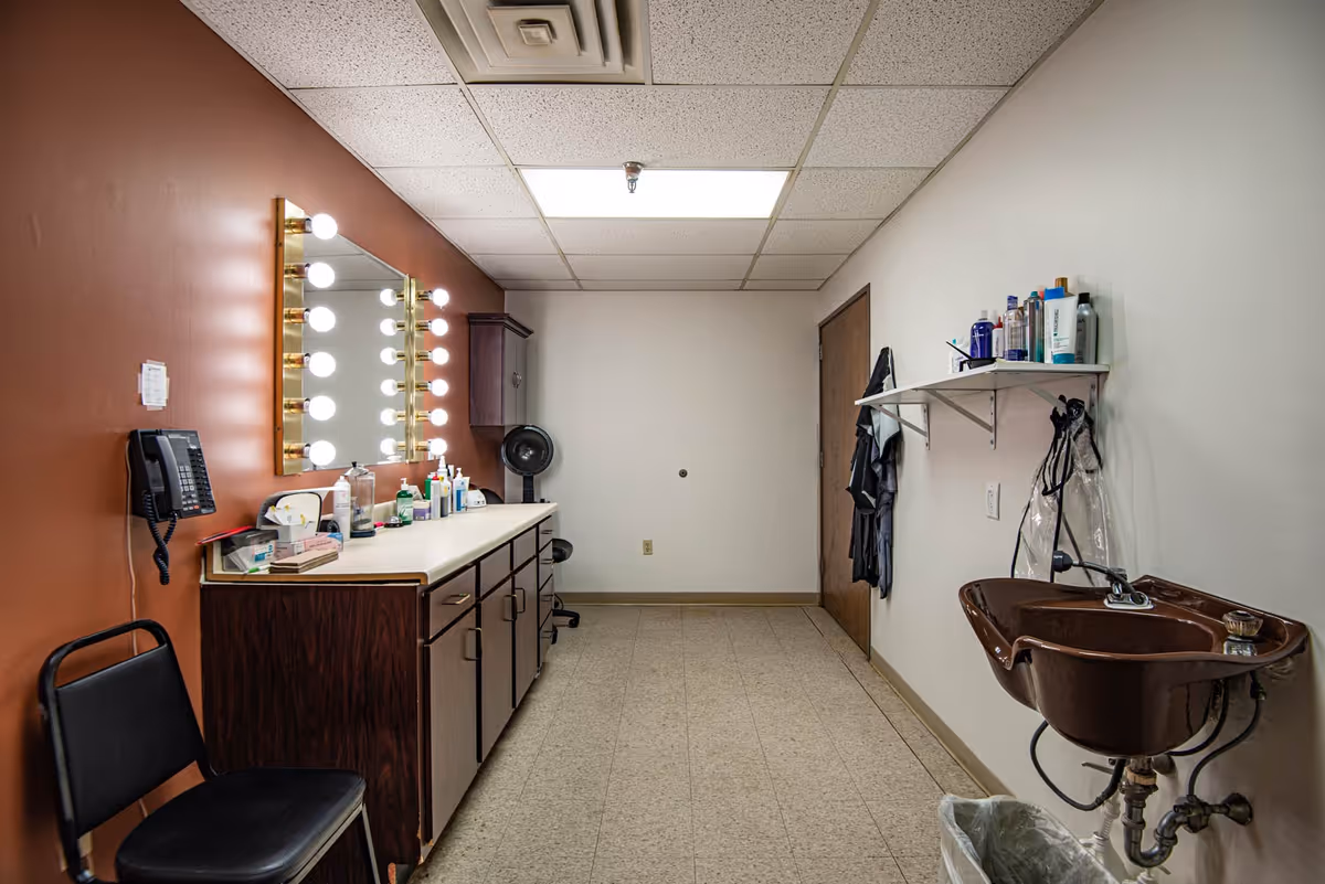 A well-lit room with a large mirror surrounded by vanity lights above a countertop with various personal care products. There is a black chair in front of the counter, a wall-mounted telephone, a brown sink on the right wall with more personal care items on a shelf above it, and a closed wooden door at the far end of the room.