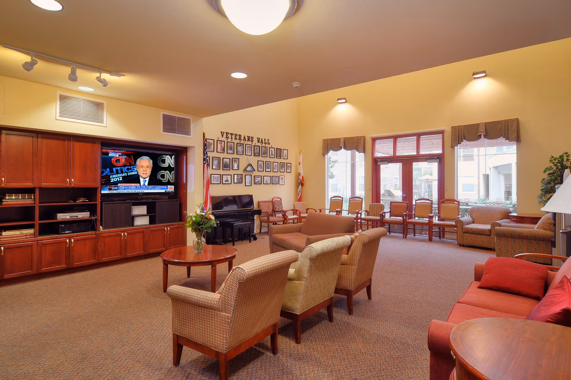 A spacious living room area with multiple chairs and sofas arranged around a central coffee table with a vase of flowers. A large wooden entertainment center with a television is on the left wall, showing a news program. The back wall features a 'Veterans Wall' with framed photos and an American flag. Large windows and glass doors let in natural light, and there is a piano in the corner.