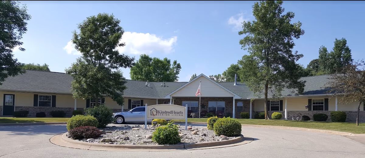 Front exterior view of KindredHearts Senior Living facility in Greenville, showing a single-story building with a green roof, beige siding, and a stone facade. There is a circular driveway with a landscaped island in the center featuring bushes and rocks, and an American flag is visible near the entrance.