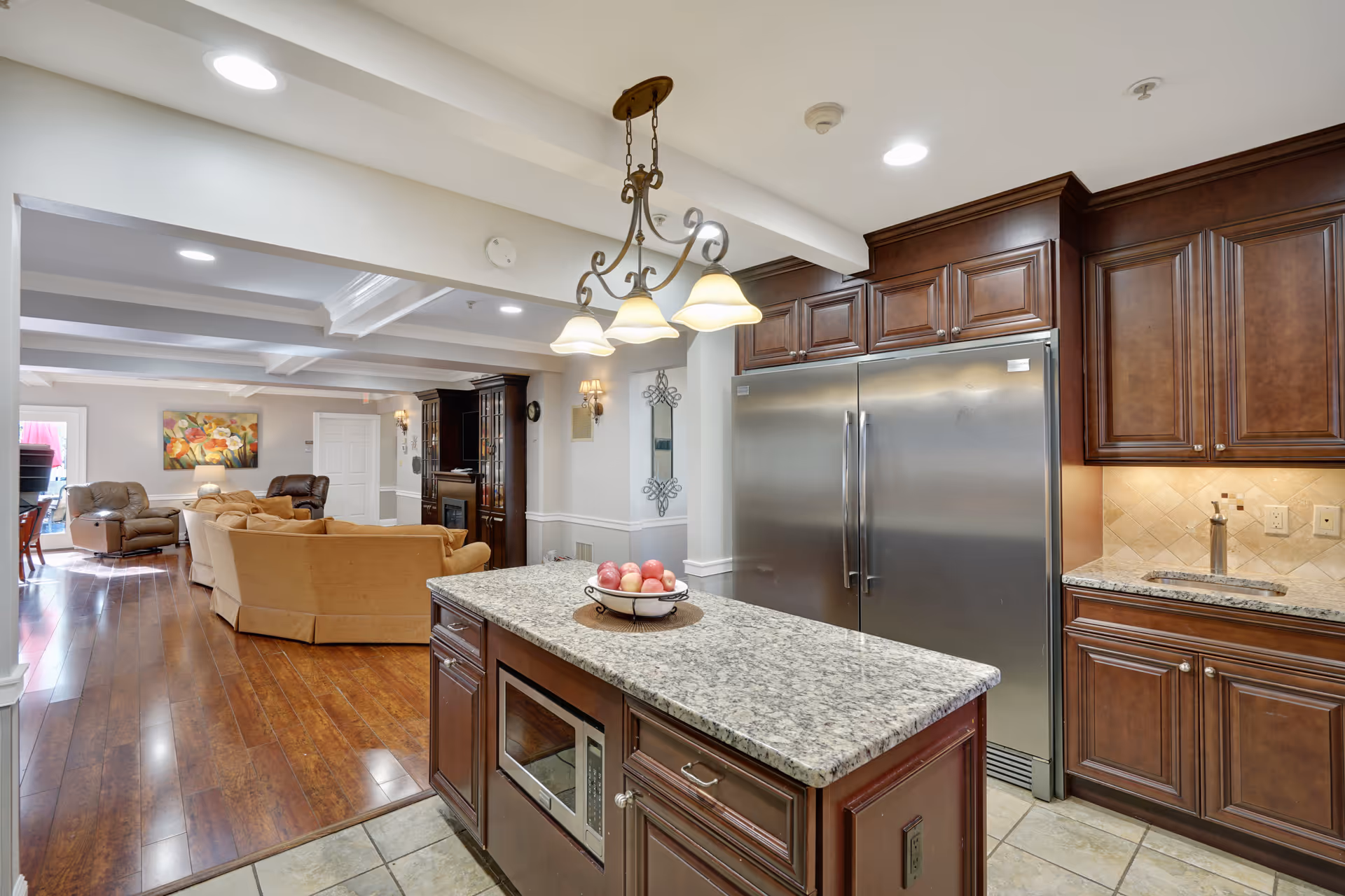 Interior view of a senior living facility showing a kitchen area with a granite countertop island, dark wood cabinets, a stainless steel refrigerator, and a microwave. The kitchen opens into a living room area with beige sofas, a recliner, a wooden floor, and a colorful painting on the wall.