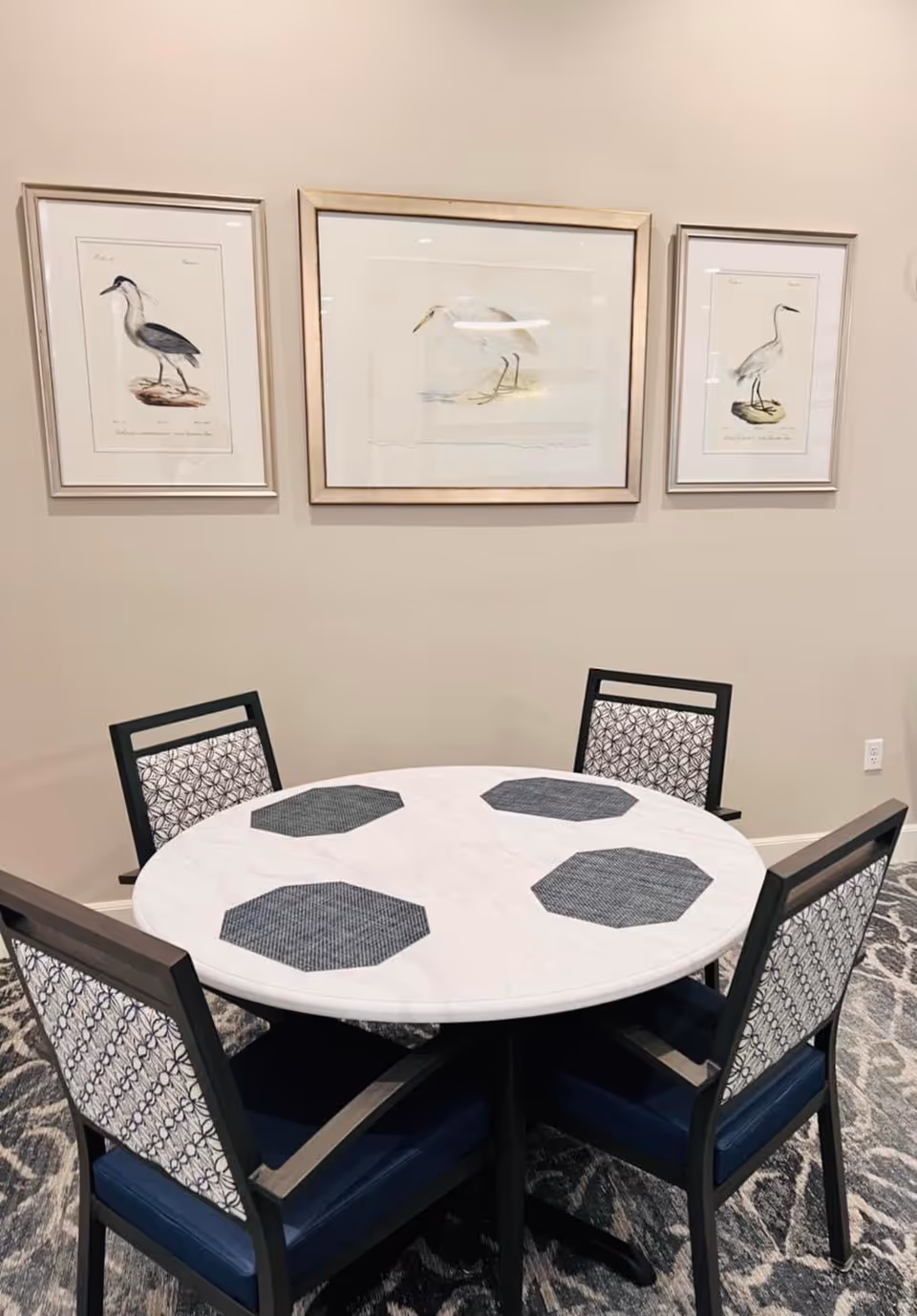 A round white table with four black chairs featuring patterned backs and blue seats. The table has four dark gray octagonal placemats. Behind the table, there are three framed bird illustrations hanging on a beige wall. The floor has a patterned carpet.
