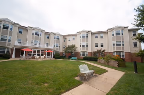 Exterior view of a three-story senior living facility building with beige and brick facade, large windows, and a well-maintained lawn. There are outdoor seating areas with red umbrellas and a paved walkway leading through the grassy courtyard.