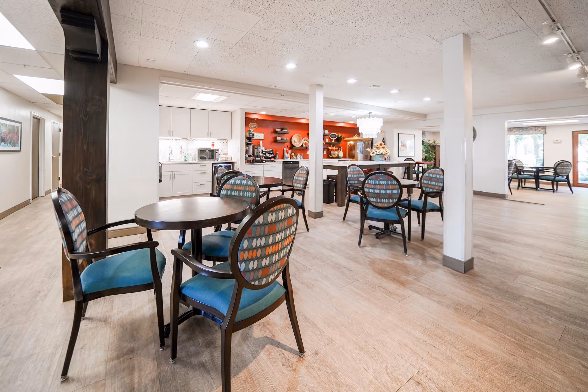 Open dining area with round tables and patterned chairs facing a kitchenette and serving counter under recessed lighting.