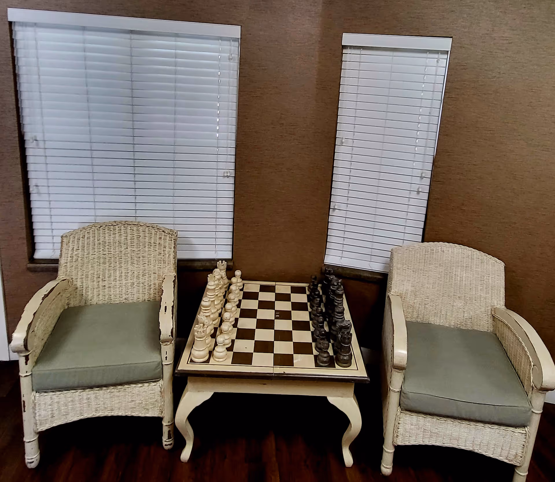 Two wicker armchairs with green cushions facing each other with a small table in between holding a chessboard set up for a game, positioned in front of two windows with white blinds in a room with brown walls and wooden floor.