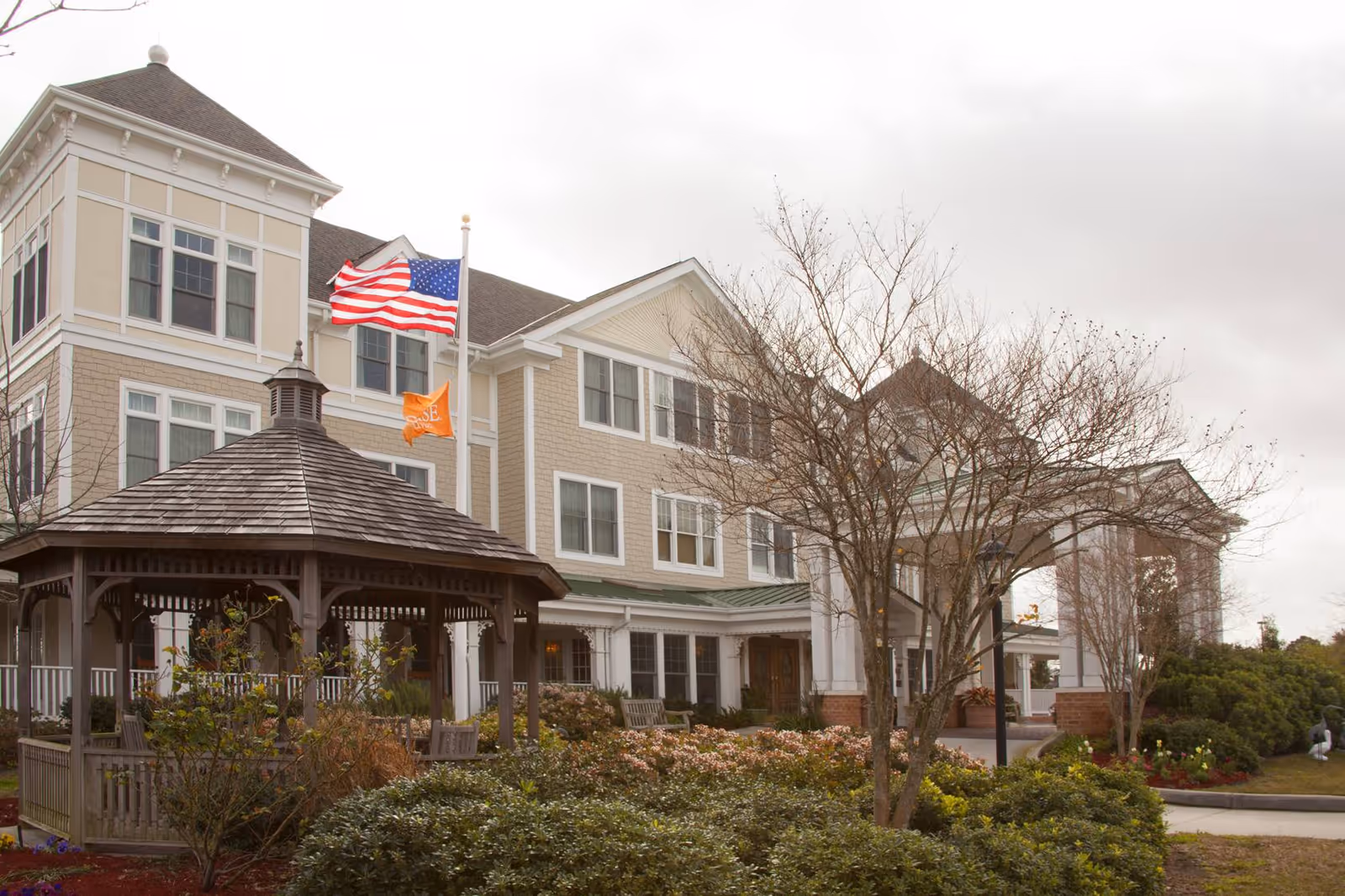Exterior view of a multi-story senior living facility with beige siding and white trim. There is a wooden gazebo in the foreground surrounded by bushes and trees, some without leaves. Two flagpoles display the American flag and an orange flag with the word 'Sunrise'. The sky is overcast.