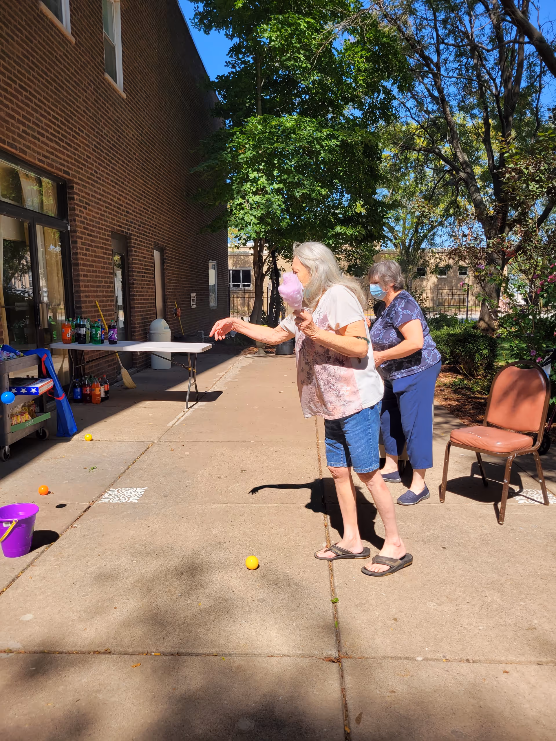 Two elderly women playing a ball toss game outside on a sunny day near a brick building. One woman is holding pink cotton candy while tossing a ball, and the other woman is standing behind her wearing a face mask. There are chairs, a table with drinks, and trees in the background.