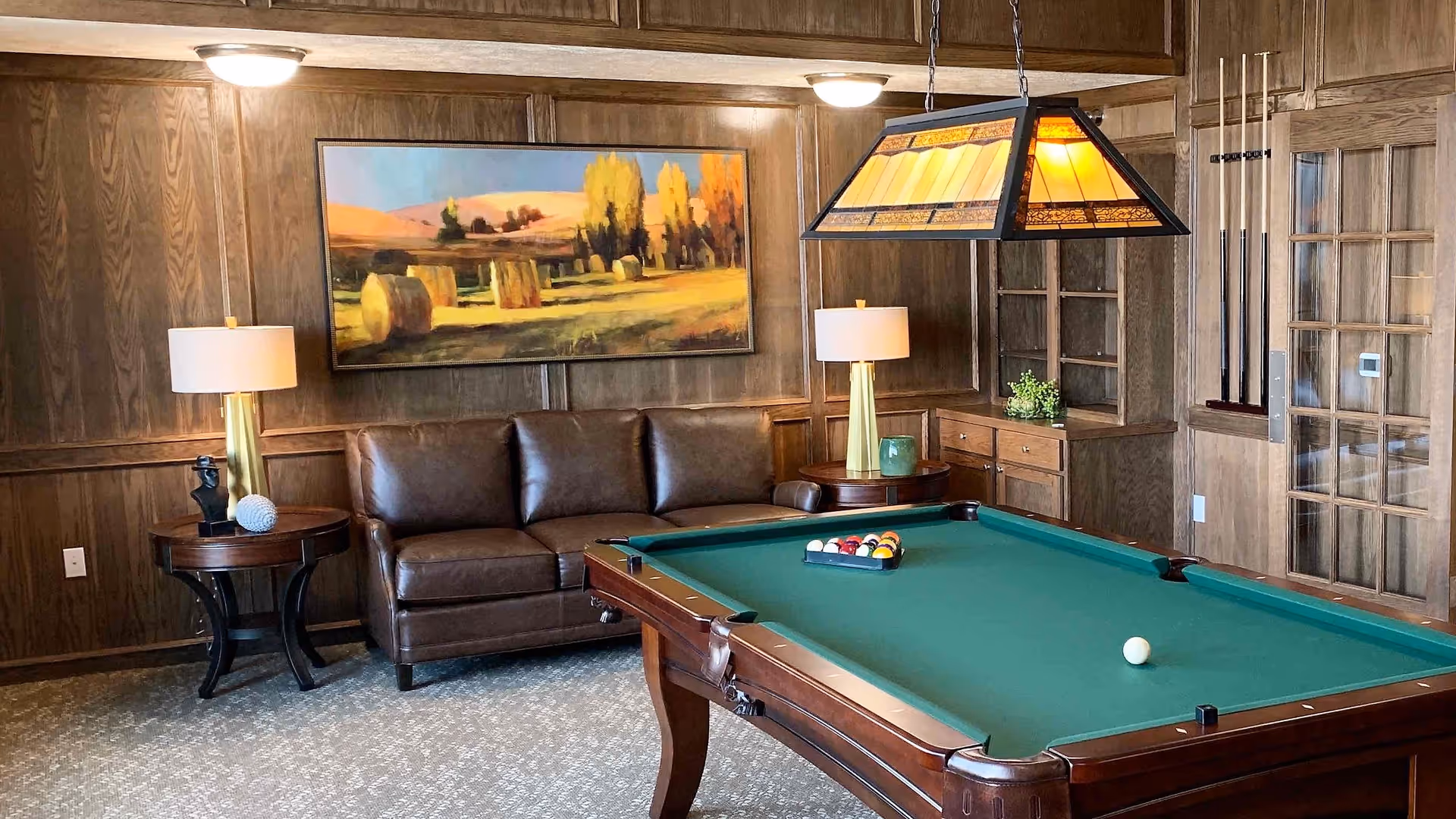 A wood-paneled lounge featuring a pool table, leather sofa, side tables with lamps, and framed landscape art on the wall.