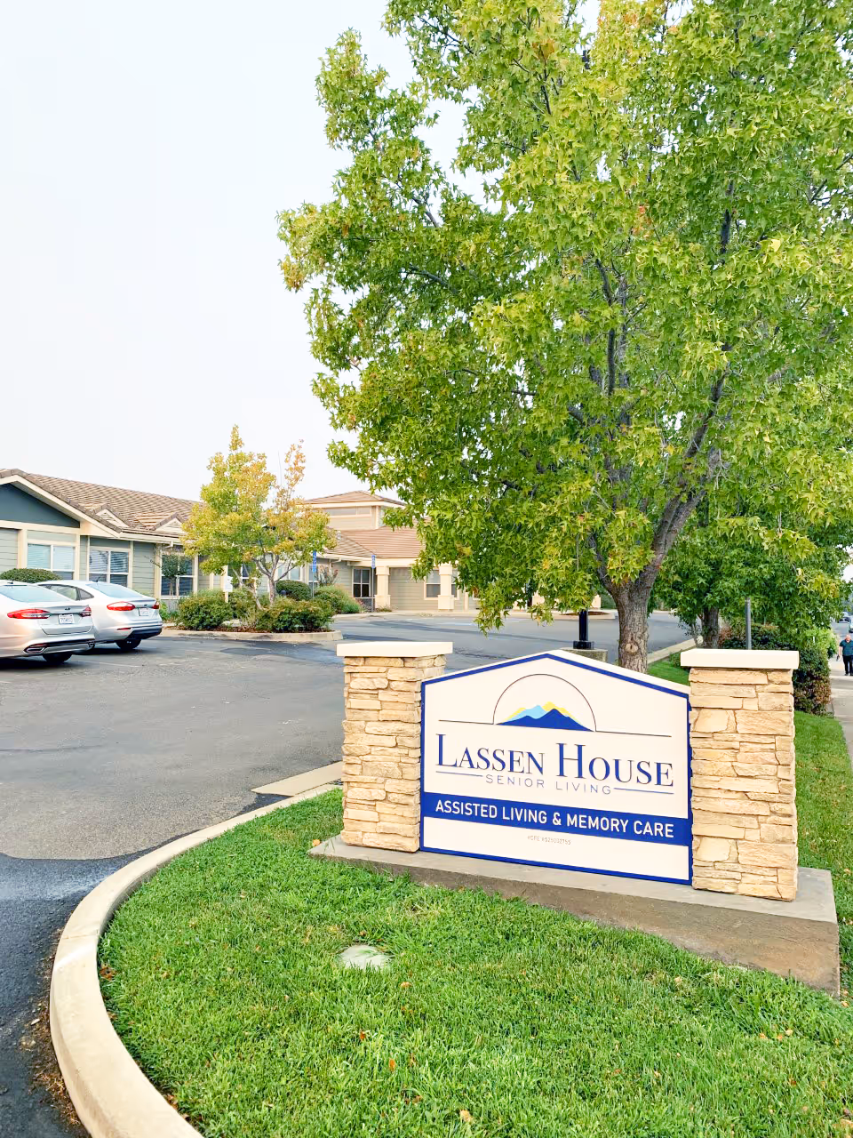 Outdoor view of Lassen House Senior Living facility sign with stone pillars on either side, green grass and trees surrounding the area, and part of the building and parked cars visible in the background.