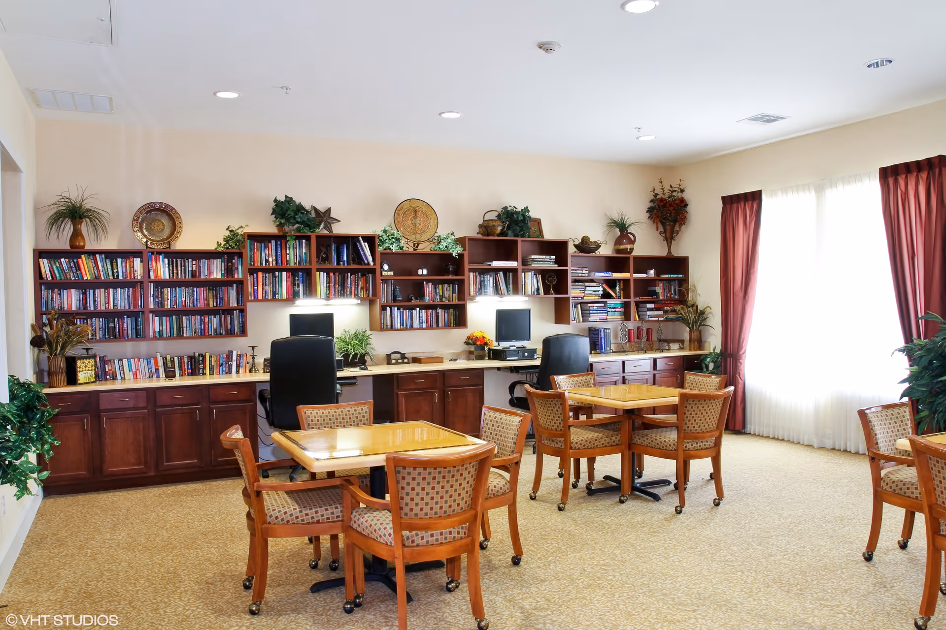 A bright room with beige walls and carpet, featuring two wooden tables each surrounded by four cushioned chairs with patterned upholstery. Along the back wall are built-in wooden shelves filled with books and decorative items, with two computer workstations and office chairs underneath. Large windows with sheer white curtains and red drapes allow natural light to fill the space. Several potted plants and decorative plates add a homey touch.