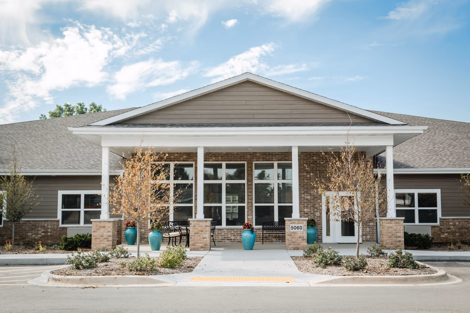 Front exterior view of a single-story building with a covered entrance supported by white columns. The building has a combination of brick and siding walls, large windows, and a gray shingled roof. There are small landscaped areas with bushes and young trees, and blue ceramic planters with flowers near the entrance. The sky is partly cloudy with blue patches.