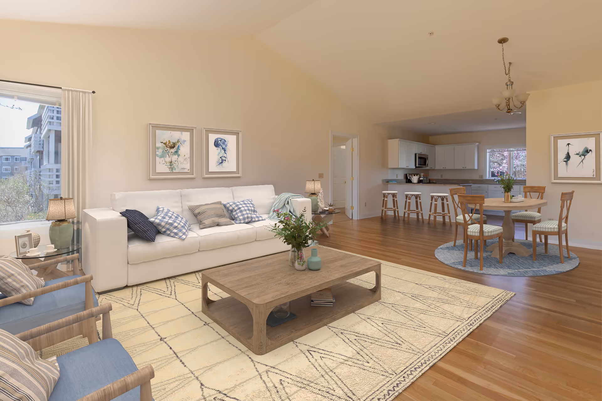 A bright and spacious living area with a white sofa adorned with multiple cushions, two blue armchairs, a wooden coffee table with decorative items, and a patterned rug. In the background, there is a dining area with a round wooden table and four chairs on a blue circular rug, and a kitchen with white cabinets, a countertop with four bar stools, and a window letting in natural light.