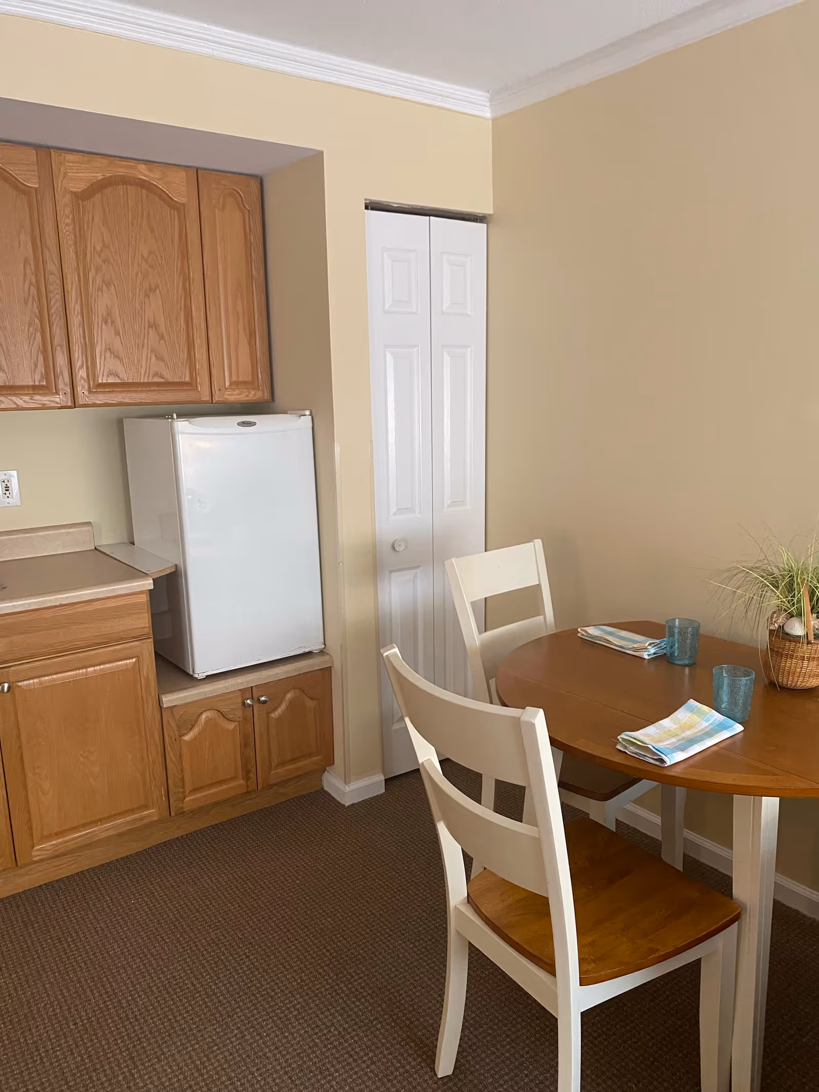 A small dining area with a round wooden table set with two folded napkins and two blue drinking glasses. Two white chairs with wooden seats are positioned around the table. To the left, there is a compact kitchen area with wooden cabinets, a small white refrigerator, and a white door closet in the background. The walls are painted beige and the floor is carpeted.
