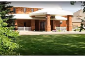 Exterior view of Derian Lodge Senior Living facility showing a brick building with a covered entrance supported by brick columns, surrounded by green lawn and trees.