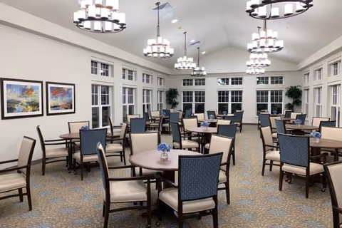 A spacious dining room with multiple round tables and chairs arranged neatly. The room features large windows along the walls, allowing natural light to enter. The ceiling is vaulted with several modern chandeliers hanging down, and there are framed landscape paintings on one wall. The carpet has a patterned design, and small flower arrangements are placed on each table.