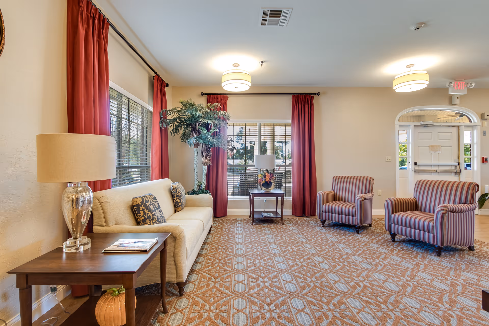 A bright and welcoming living room area with a beige sofa adorned with patterned cushions, two striped armchairs, a wooden side table with a lamp and magazines, and a small table with a lamp and flower arrangement. The room features large windows with red curtains, a patterned carpet, and ceiling lights. A potted plant is placed near the window, and an exit door is visible in the background.