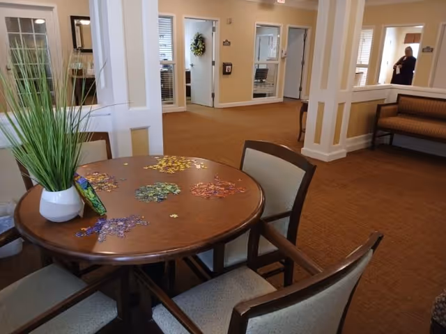 Round table with chairs and a partially assembled jigsaw puzzle in a senior living common area with seating and a hallway in the background.