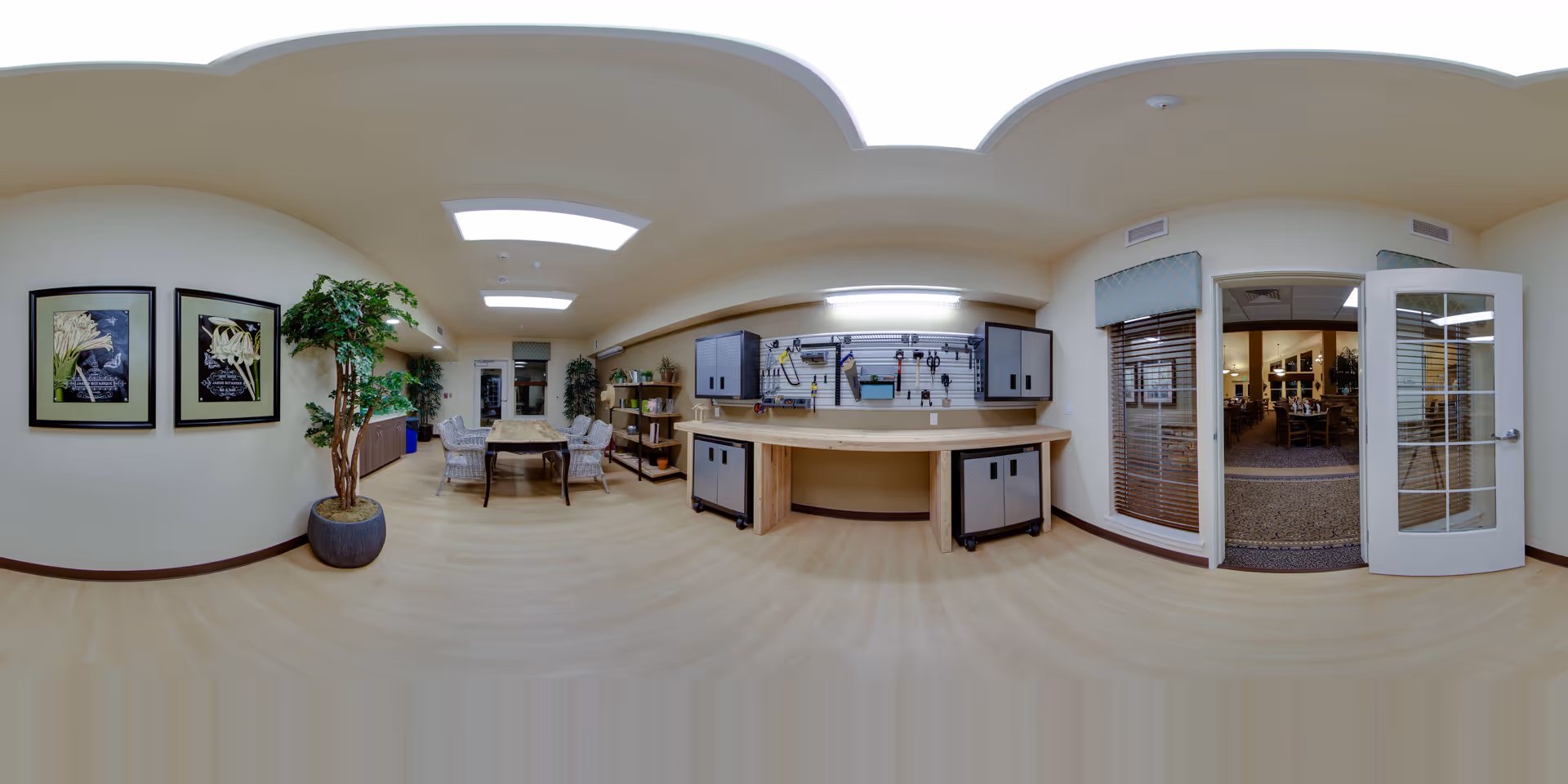 Bright interior common activity room with a long workbench and tool storage, a table with chairs, potted plants, and a door opening to a dining area.