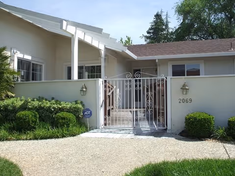 Exterior view of a single-story residential building with a white metal gate and a small enclosed front patio. The building has beige walls, a brown roof, and two wall-mounted lantern-style lights on either side of the gate. The address number 2069 is visible on the right side of the gate. There are green shrubs and grass in front of the building, and trees are visible in the background under a clear sky.