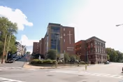 Exterior view of Marian Manor Nursing Home, a multi-story brick building with a glass entrance and a cross mounted on the front. The building is situated at a street corner with sidewalks and some greenery around the entrance.