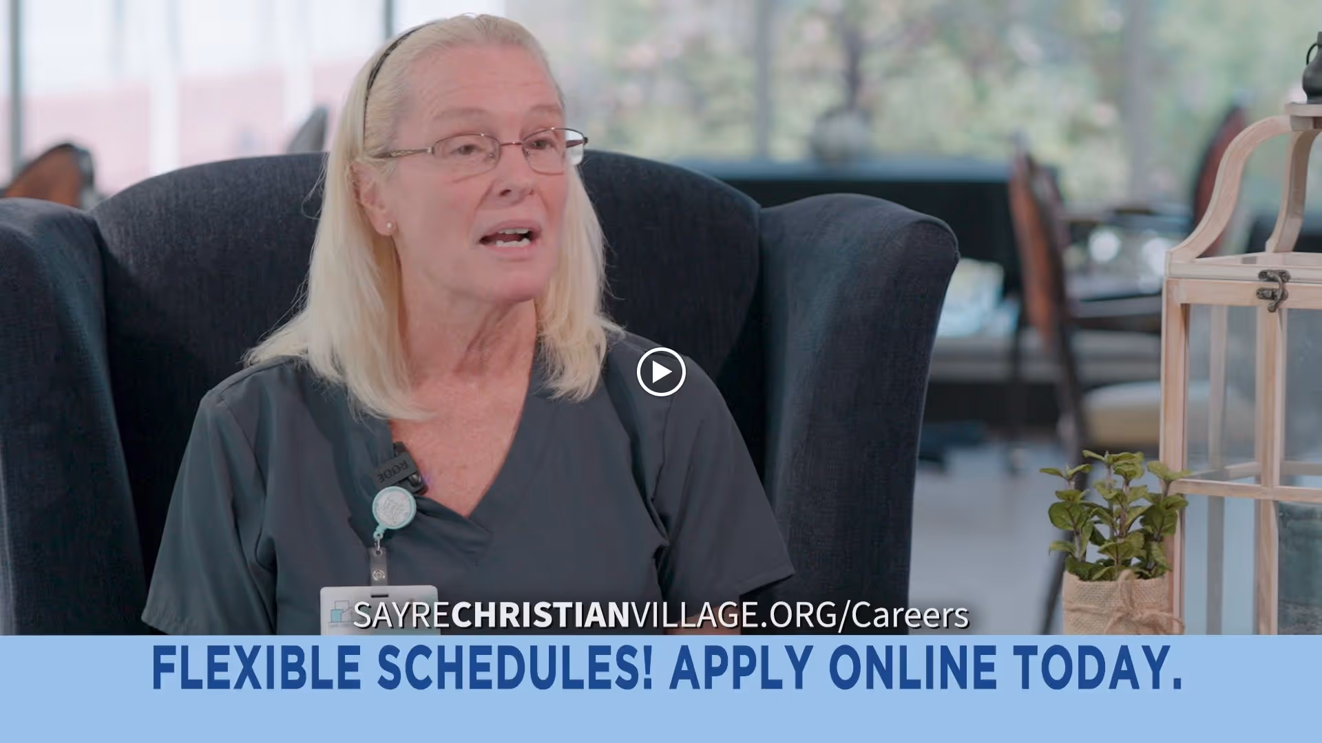 A woman with blonde hair and glasses wearing dark scrubs is sitting in a dark armchair, speaking. Behind her is a bright room with large windows and some furniture. There is a small plant and a decorative lantern on a table to the right.