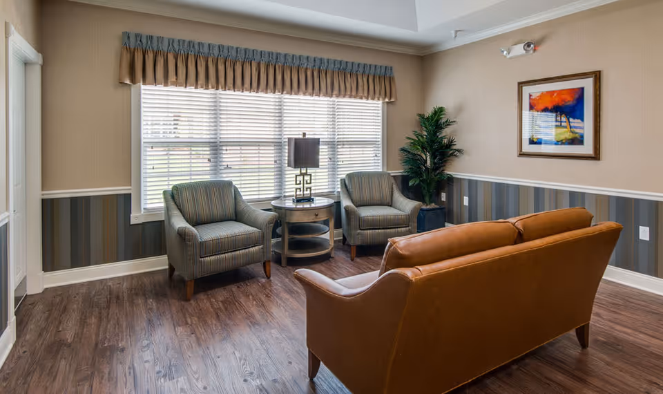 A cozy sitting area in a senior living facility with two striped armchairs and a brown leather sofa arranged around a round wooden side table with a lamp. The room has wood flooring, a large window with blinds and a valance, a potted plant in the corner, and a colorful framed painting on the wall.