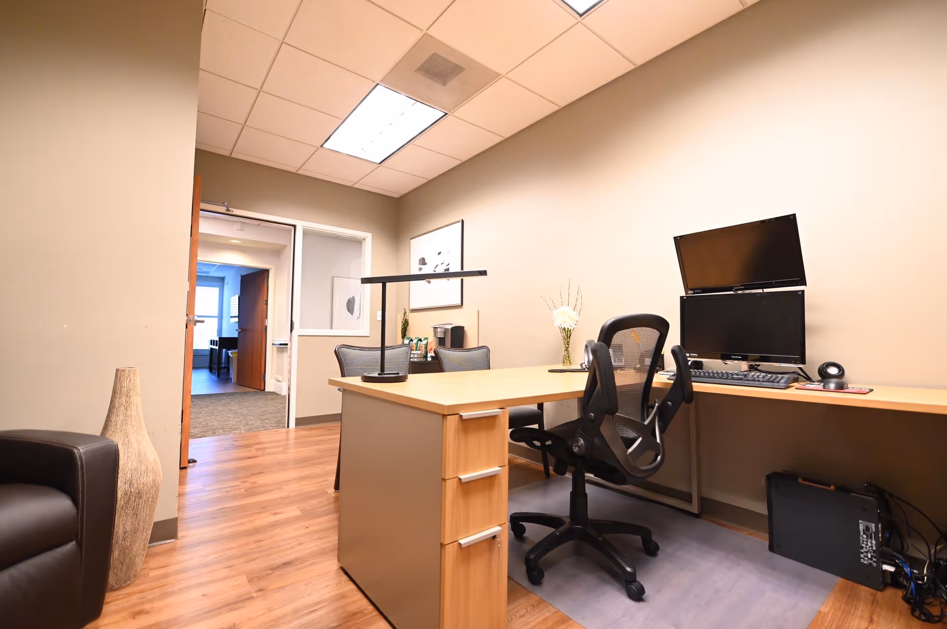 An office space with a wooden desk, black office chair, dual computer monitors, keyboard, and a desk lamp. The room has beige walls, wood flooring, and a doorway leading to another room with carpet. There is a black leather chair and a tall decorative vase in the corner.
