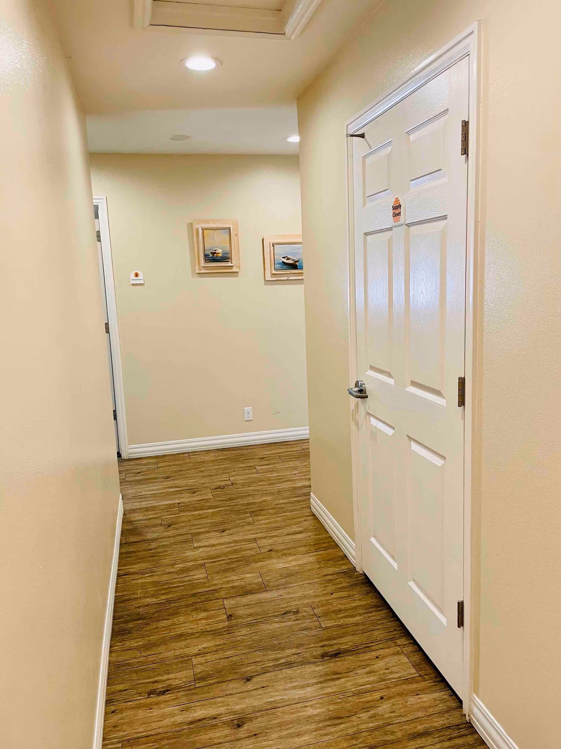 A narrow hallway with light beige walls and wood-patterned flooring. There is a white door on the right side with a small sign that reads 'Storage Closet.' Two framed pictures of boats are hanging on the far wall. The hallway is lit by recessed ceiling lights.
