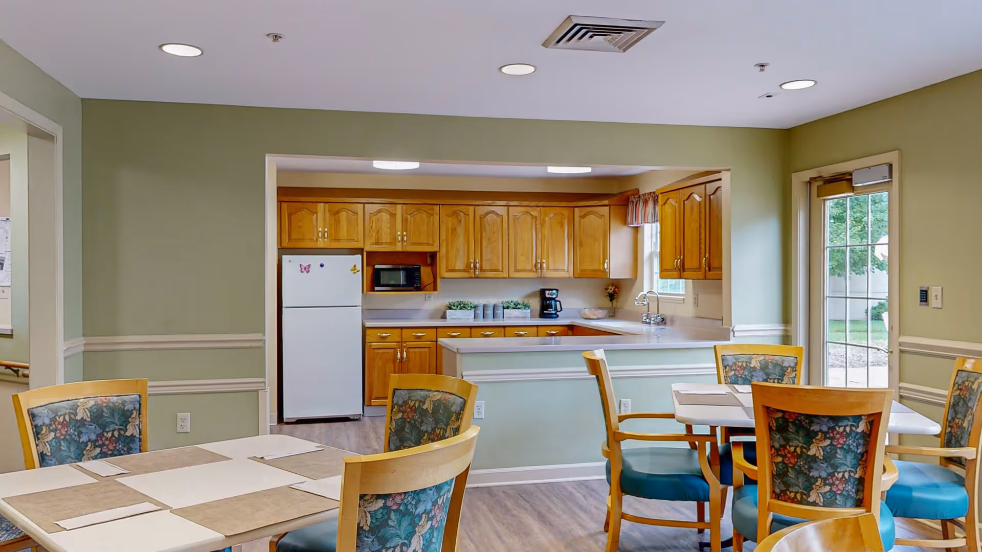 A dining area with two tables, each surrounded by wooden chairs with floral patterned cushions. The room has light green walls with white trim and wood flooring. In the background, there is a kitchen with wooden cabinets, a white refrigerator, a microwave, a coffee maker, and a window with a small curtain. A glass door on the right side leads outside to a garden area.