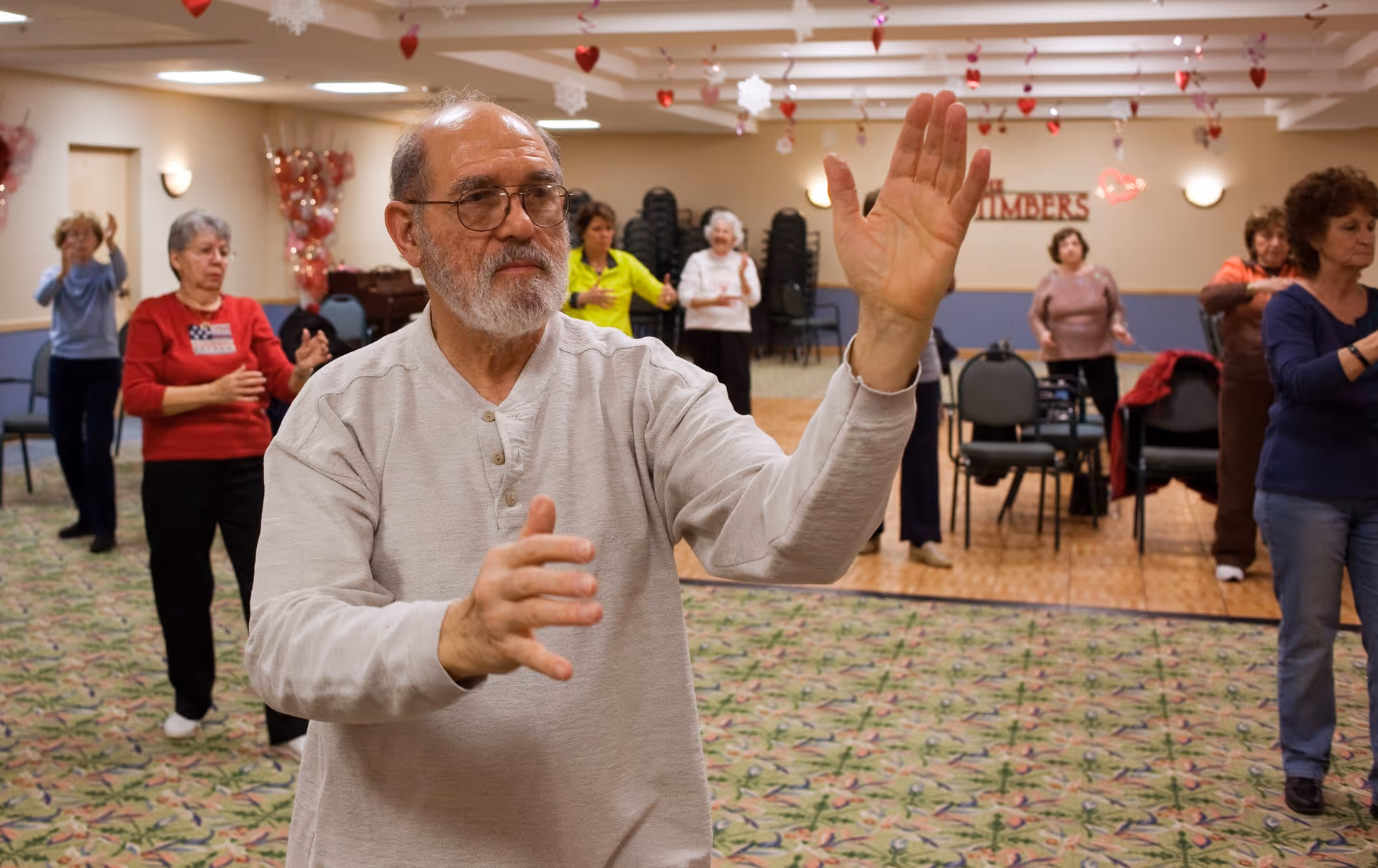 A group of elderly people participating in a group exercise or tai chi session in a decorated community room with heart and snowflake hanging decorations. The focus is on an elderly man with glasses and a beard in the foreground, with several women exercising in the background.
