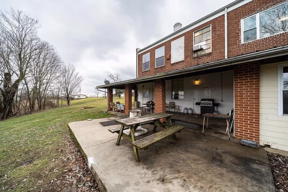 Outdoor patio area of a brick building with a wooden picnic table, several chairs, a barbecue grill, and a concrete floor. The patio is covered by a roof supported by brick columns. Surrounding the patio is a grassy area with leafless trees under a cloudy sky.