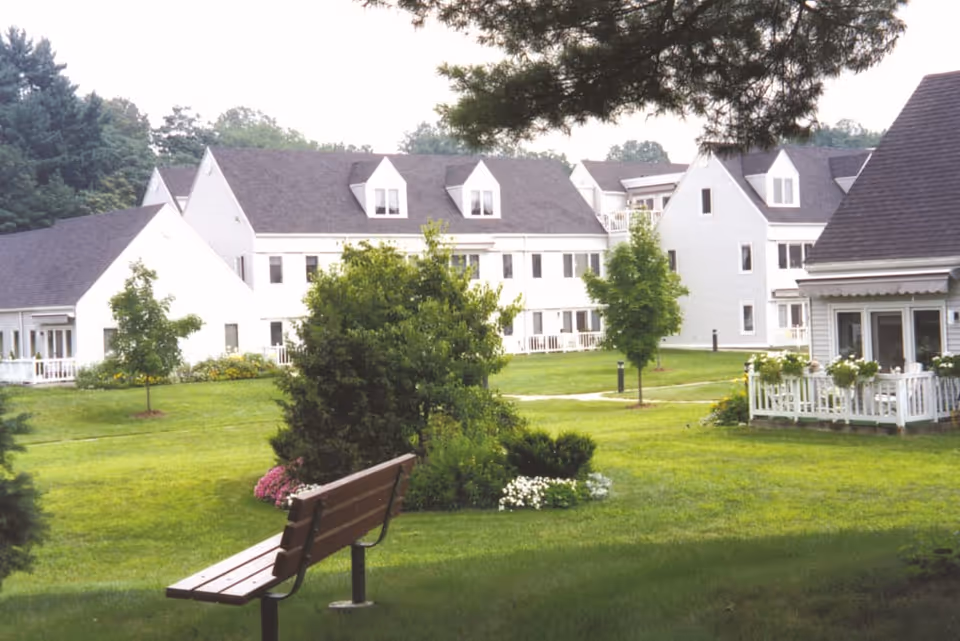 A peaceful outdoor garden area at Pomperaug Woods featuring a wooden bench, green grass, trees, bushes, and white residential buildings in the background under a partly cloudy sky.
