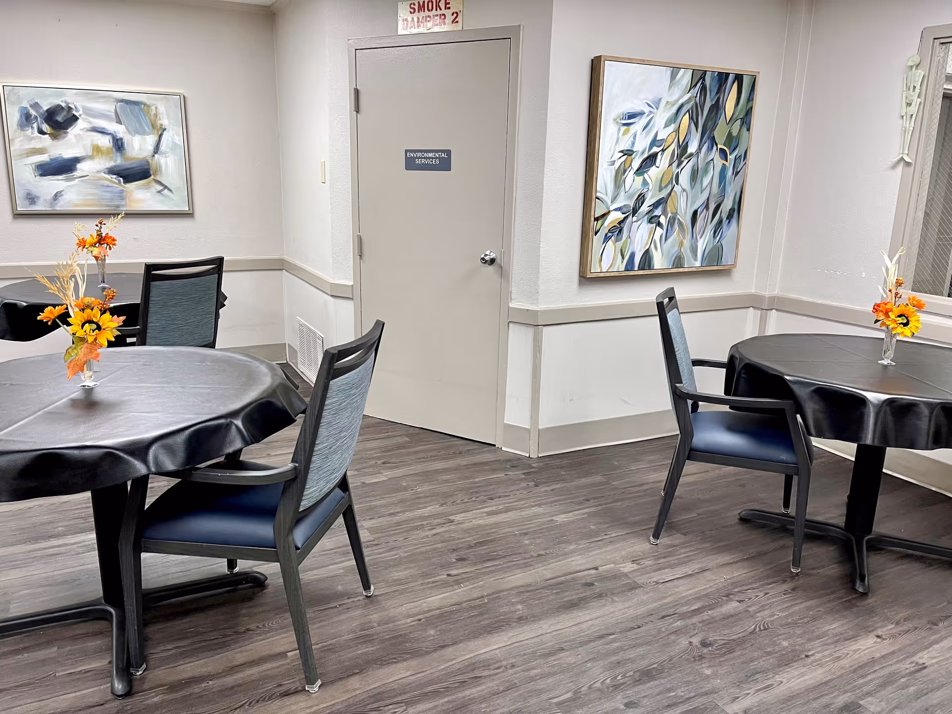 Interior view of a dining area with two round tables covered with black tablecloths, each with a small vase of artificial flowers. There are four chairs with blue cushions around the tables. The room has wood flooring, light-colored walls, two abstract paintings on the walls, and a door labeled 'Environmental Services'.