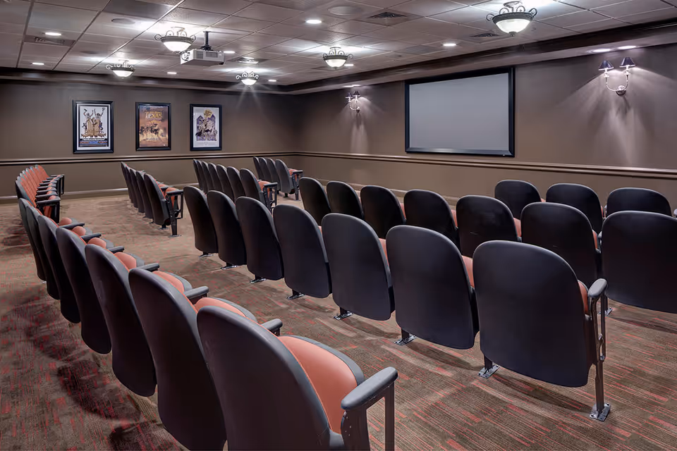 A small auditorium or screening room with several rows of theater-style seats facing a large blank projection screen on the wall. The room has brown walls, carpeted floor, framed posters on one wall, and ceiling lights.