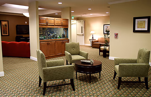 A senior living facility lounge with four green armchairs around a round coffee table, a built-in aquarium, and additional seating in the background.