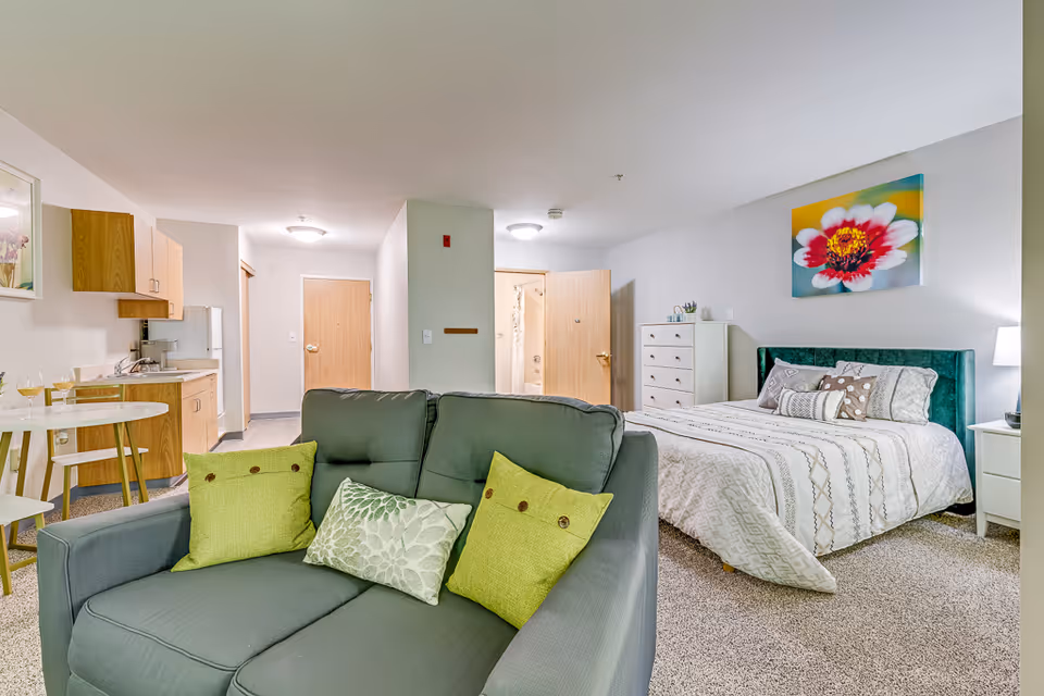 A senior living facility studio apartment featuring a green loveseat with three decorative pillows in the foreground. Behind the loveseat is a bed with a green upholstered headboard, white and patterned bedding, and a nightstand with a lamp. To the left, there is a small dining table with two chairs and a kitchenette with wooden cabinets, a sink, and a refrigerator. Two doors are visible in the background, one leading to a bathroom. The walls are light-colored, and a colorful flower painting hangs above the bed.