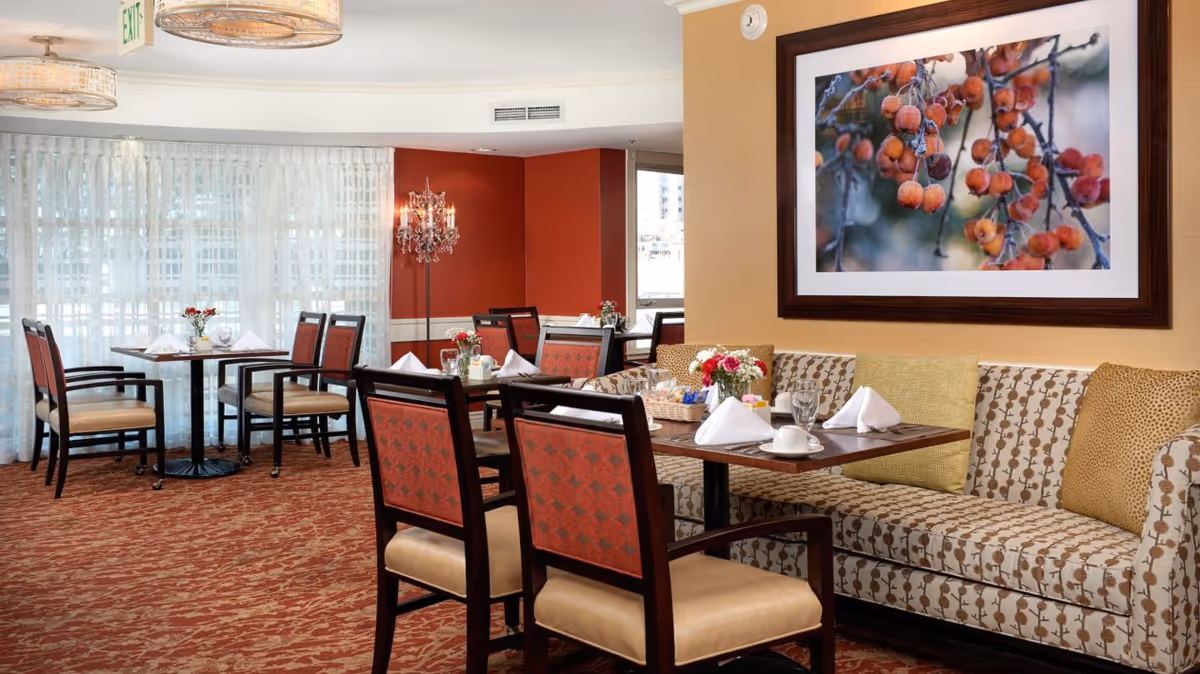 Comfortable dining room with set tables, wooden chairs, patterned banquette seating and framed artwork on a warm-colored wall.