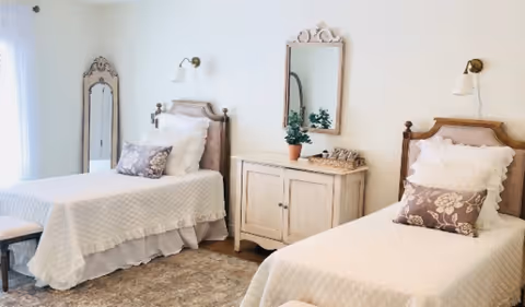 A bright bedroom with two twin beds featuring white quilted bedspreads and decorative pillows. Between the beds is a cream-colored cabinet with a potted plant and a tray on top, and a decorative mirror hanging above it. A tall, ornate standing mirror is positioned in the corner near a window with sheer white curtains. The room has a soft, neutral color palette and a cozy rug on the floor.