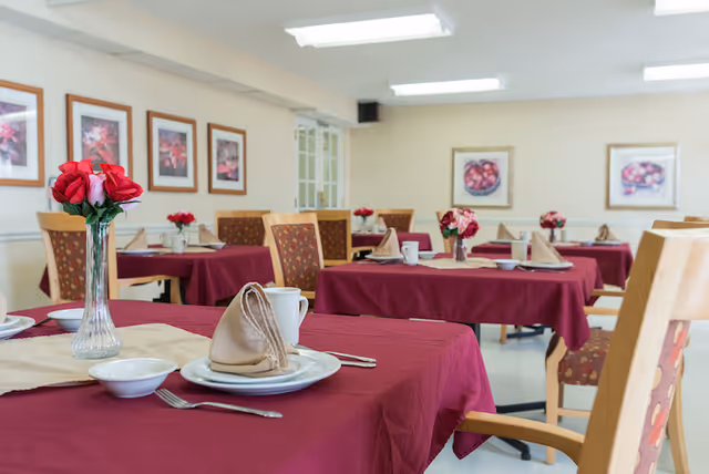 Dining room with multiple tables covered in burgundy tablecloths, set with plates, napkins, mugs, and flower centerpieces.