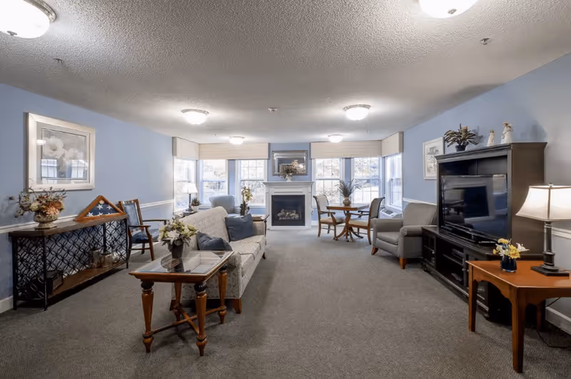 A cozy living room with light blue walls, a gray carpet, and multiple seating options including a sofa, armchairs, and wooden chairs. There is a fireplace centered on the far wall beneath a framed picture, flanked by large windows with blinds. A wooden TV stand with a flat-screen television and a side table with a lamp and flowers are on the right side. On the left side, there is a console table with decorative items and a framed picture above it. The room is softly lit by ceiling lights and a table lamp.
